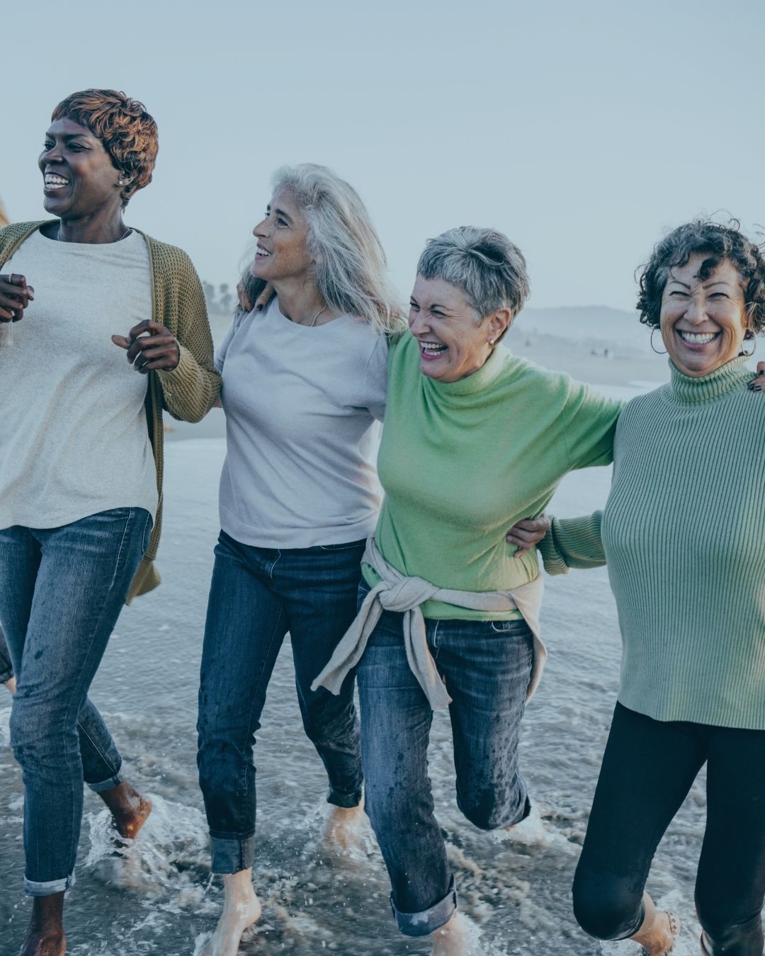 A group of women diverse in age, race, and physical form, walk through shallow beach waves with arms around each other, they look happy